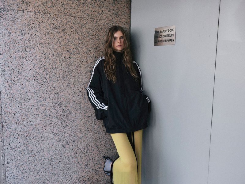 A young woman in a black Adidas jacket with white stripes poses against a grey wall and a fire safety door. Fashion editorial.