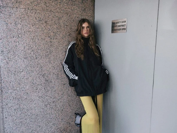 A young woman in a black Adidas jacket with white stripes poses against a grey wall and a fire safety door. Fashion editorial.