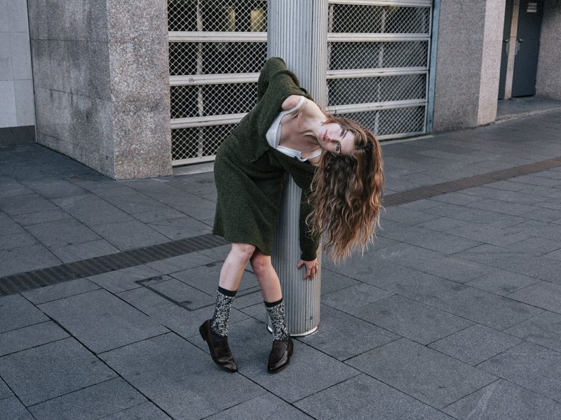 Woman in a green dress leans against a metal pole on an urban street, showcasing a fashion-forward and slightly unconventional pose.