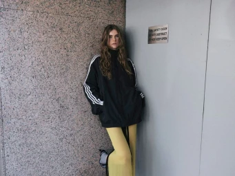 Model Anna Robinson leaning against a granite wall wearing a black track jacket and yellow trousers for the Side-note project No Standing.
