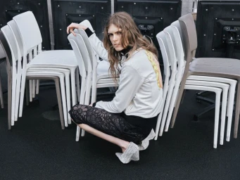 Model crouching among stacked plastic chairs in a white jacket and black lace skirt for the Side-note project No Standing