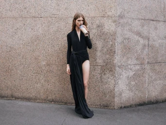 Model wearing a black draped dress drinking from a cup against a stone wall