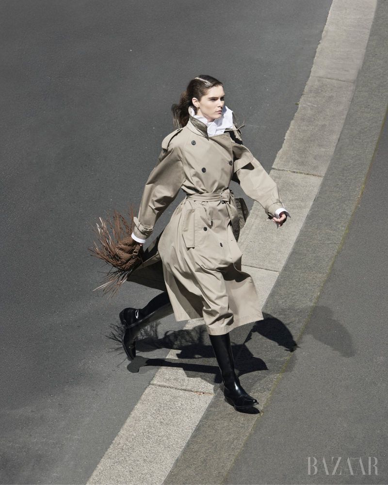 A fashion model strides confidently on a city street in a tan trench coat, black boots, and holds foliage, captured from a high angle.