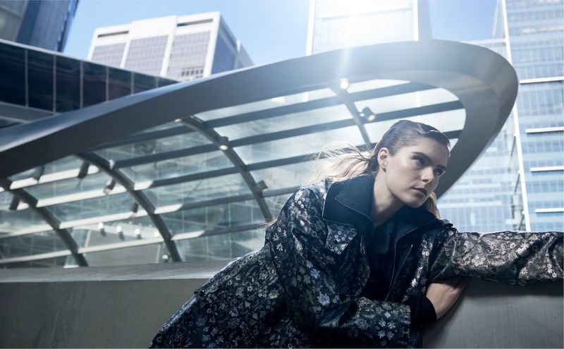 Adult female in a patterned jacket poses against a modern urban background with skyscrapers and a glass-covered walkway in daylight.