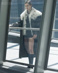 Model Anna Robinson wearing a long fur-trimmed coat and knee-high socks standing behind glass for Harper's Bazaar Australia.
