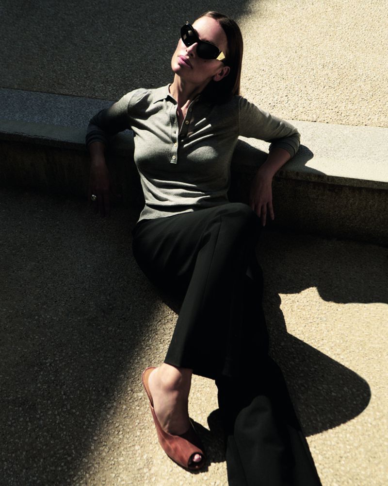 Woman in sunglasses poses outdoors in sunlight wearing a grey polo shirt, black pants, and brown mule shoes with shadows on the ground.