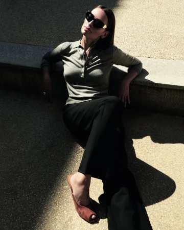 Model Almudena Fernandez wearing dark sunglasses and a grey polo top sitting outdoors in strong sunlight.