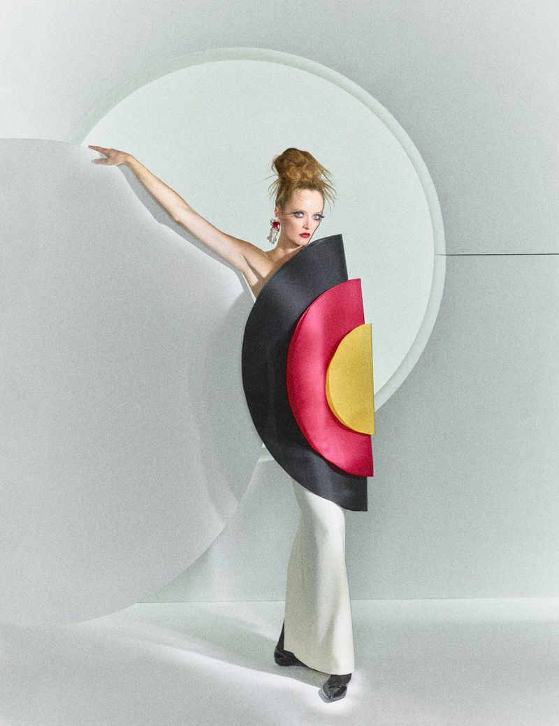 Fashion model in a white dress with geometric black, red, and yellow circular panels poses against a white studio backdrop.
