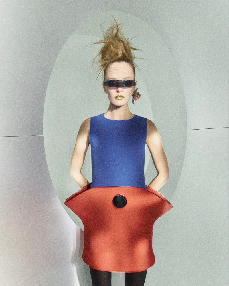 A fashion model with bold makeup wears a blue and orange outfit and dark sunglasses, posing against an illuminated white background.