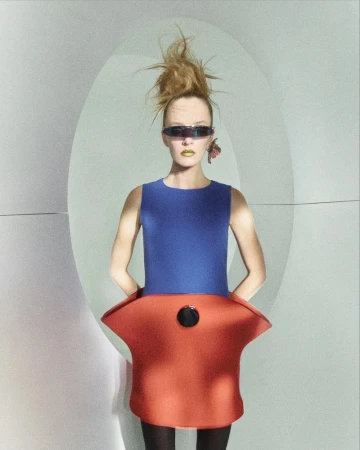 A fashion model with bold makeup wears a blue and orange outfit and dark sunglasses, posing against an illuminated white background.