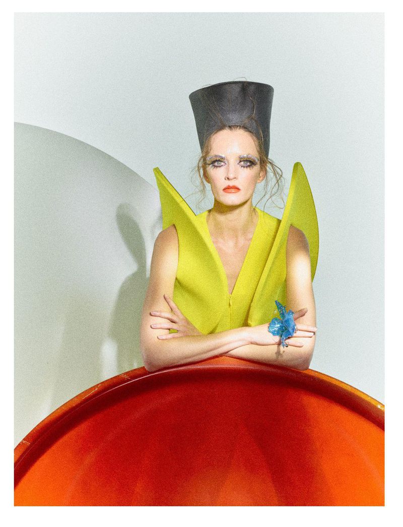 High-fashion portrait of a blonde woman wearing an avant-garde hat and yellow dress, posed in front of a white and red geometric set.