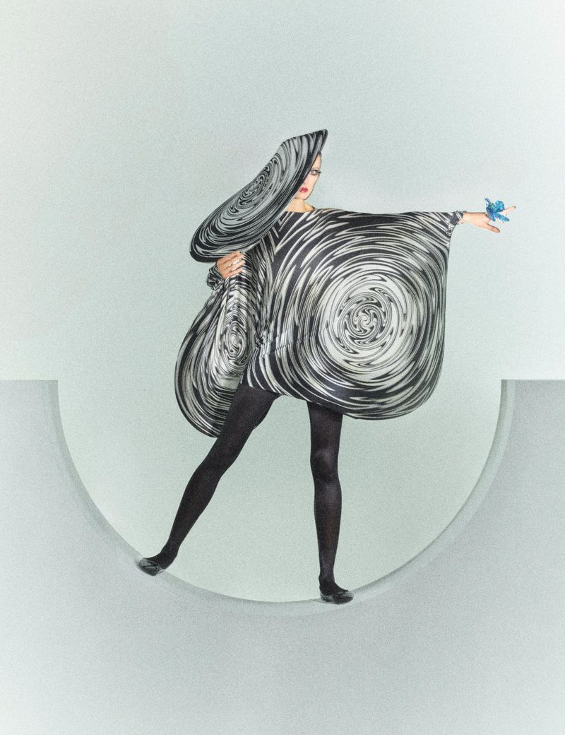 Fashion model in a black and white swirl dress and headpiece strikes a pose against a pale gray background, displaying a small bird.