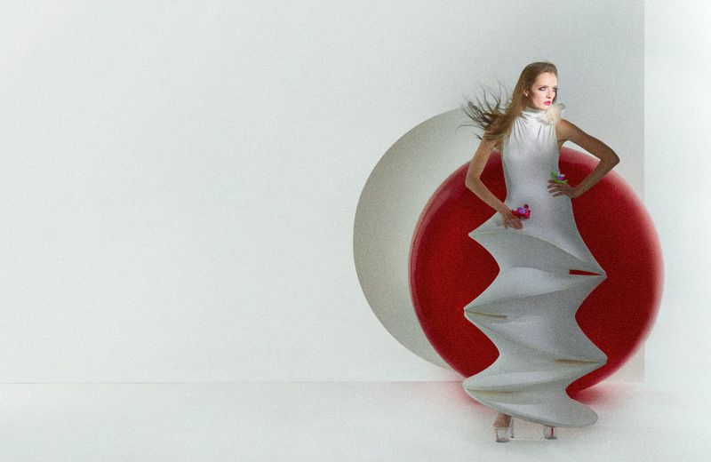 Fashion editorial shot of a woman posing in a white, sculpted dress with a red sphere in the background, set in a white studio.