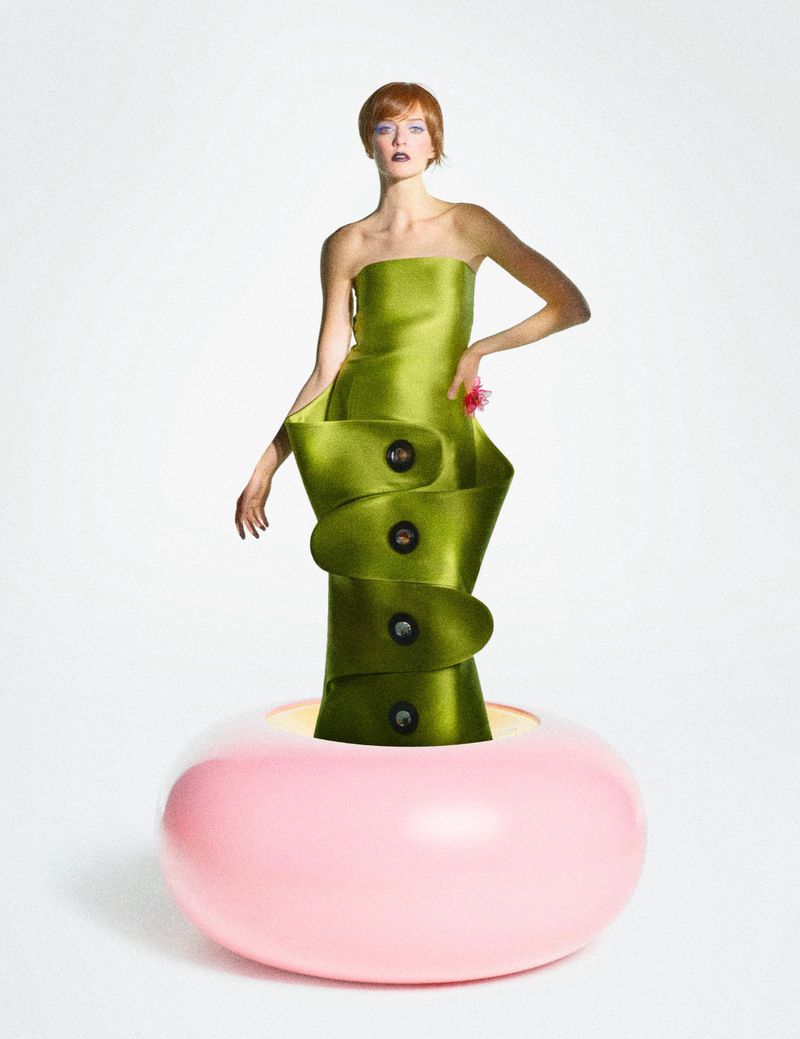 Fashion shot of a model wearing a green strapless dress with oversized buttons standing on a pink donut shaped prop against a white background.