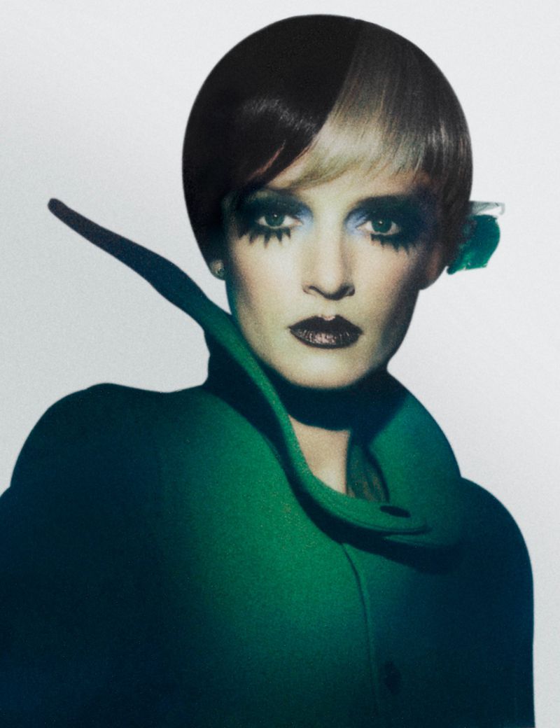Portrait of a woman with distinctive makeup and a green coat, highlighted by strategic lighting against a neutral background.