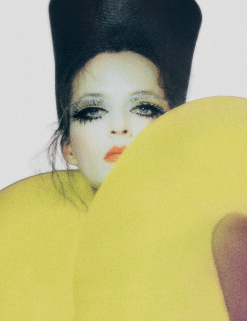 Woman with heavy eye makeup and a tall black beehive hairstyle poses with a large, round, yellow donut-shaped prop.