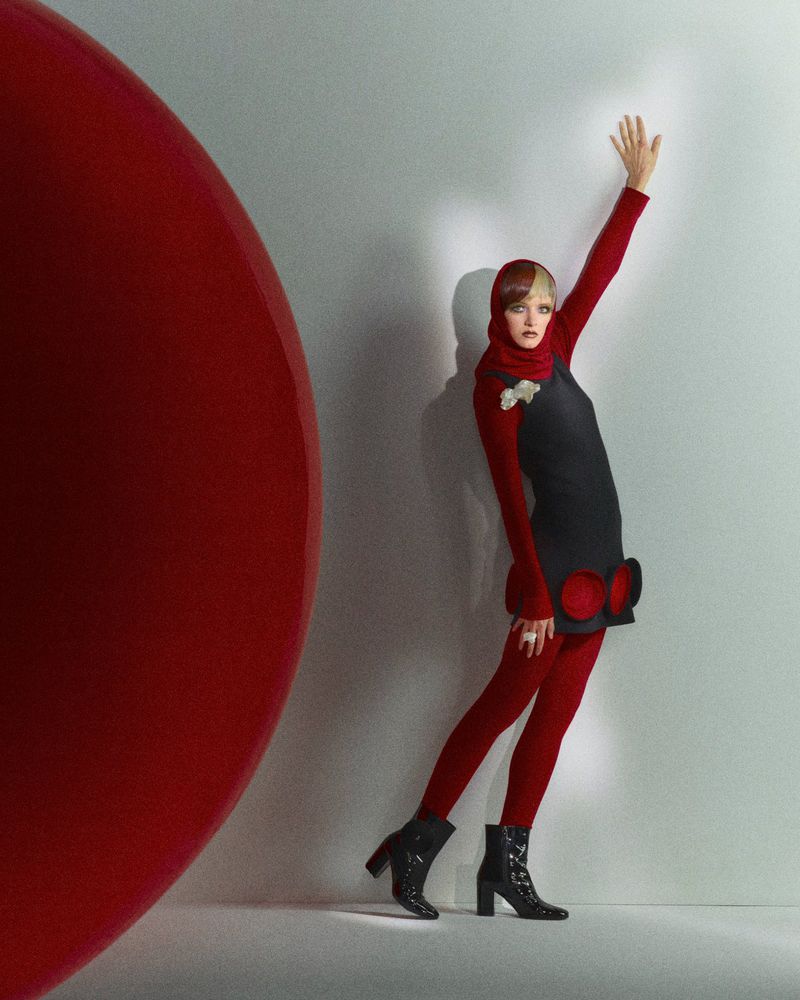 A female model poses in red tights and a black dress with a large red spherical object in a photo studio setting, fashion photography.