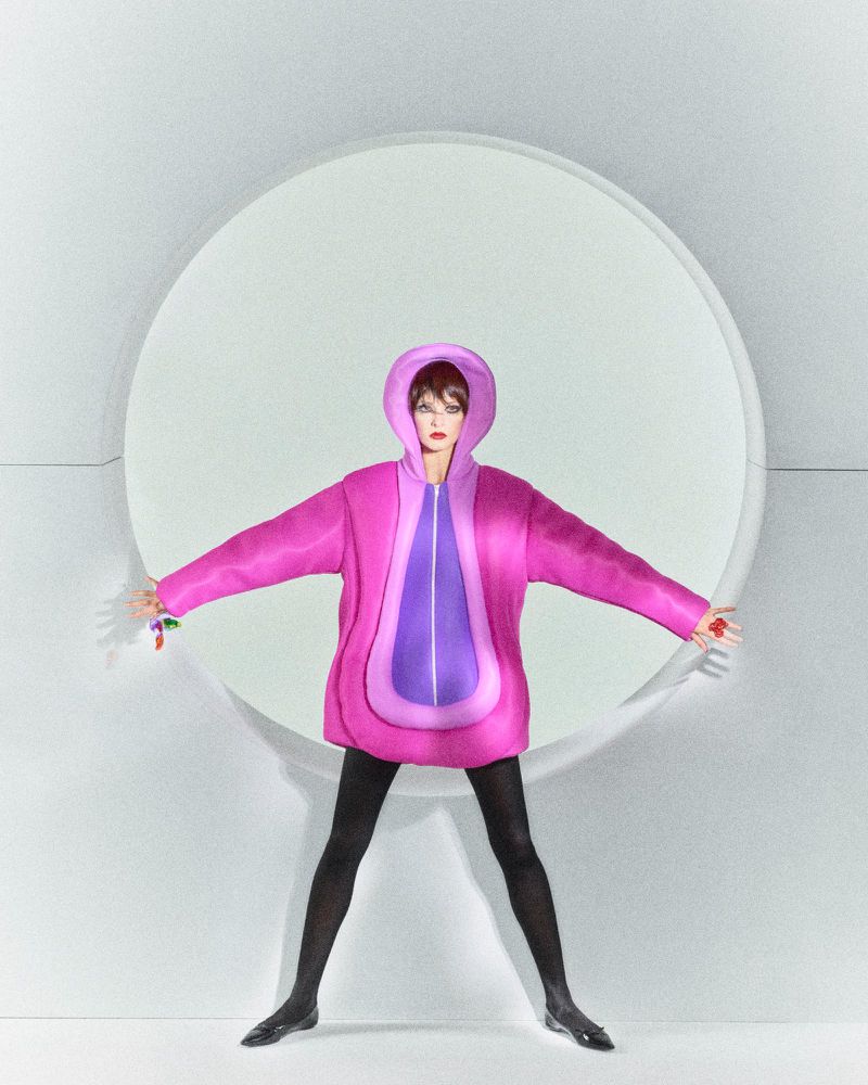 A slender adult female model wears a hooded pink and purple coat with black tights and black shoes in a minimalist, light studio setting.