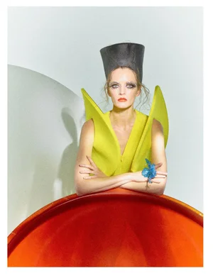 Daria Strokous wearing a lime green winged architectural top and tall cylindrical headdress, leaning over a red curved structure.