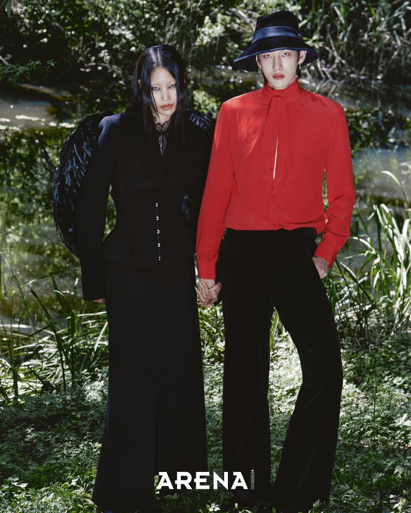 Two models standing outdoors by water, one wearing a black outfit with wings and the other in a red shirt and black hat