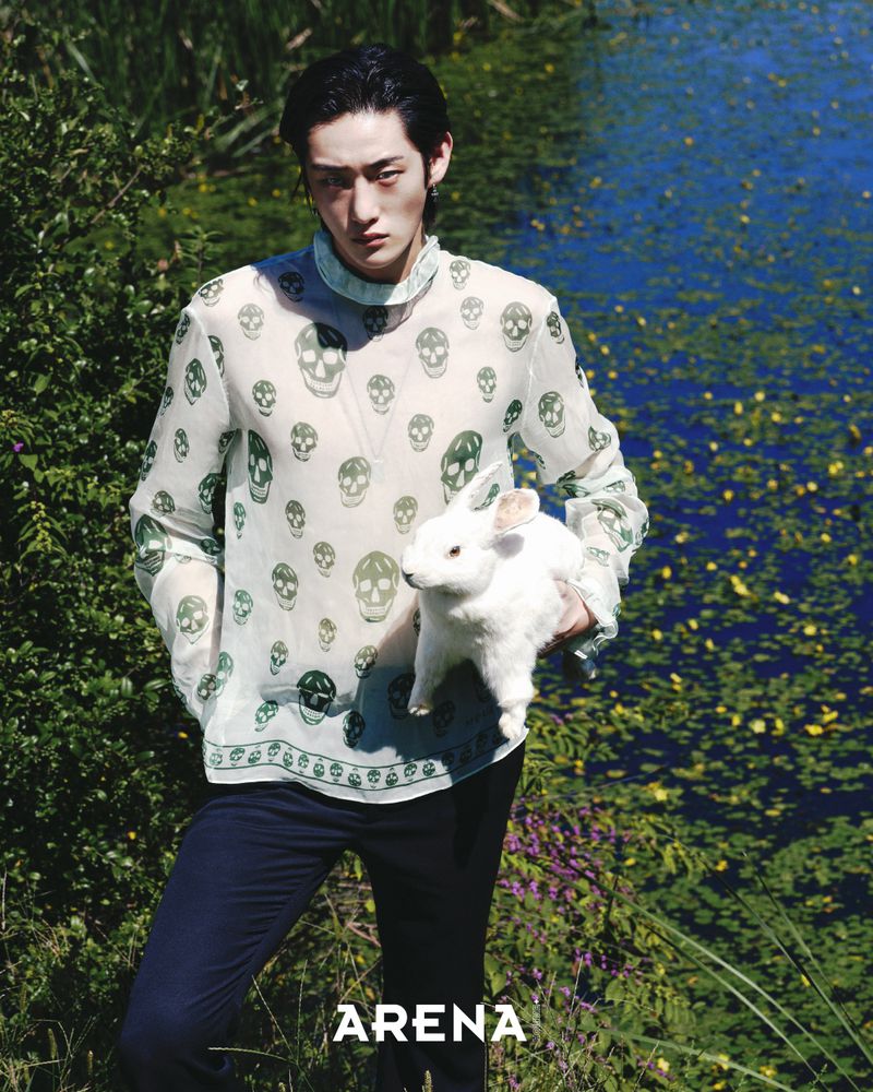 Asian man posing in nature with a white rabbit, wearing a translucent shirt with skull pattern and dark trousers, in a natural setting.