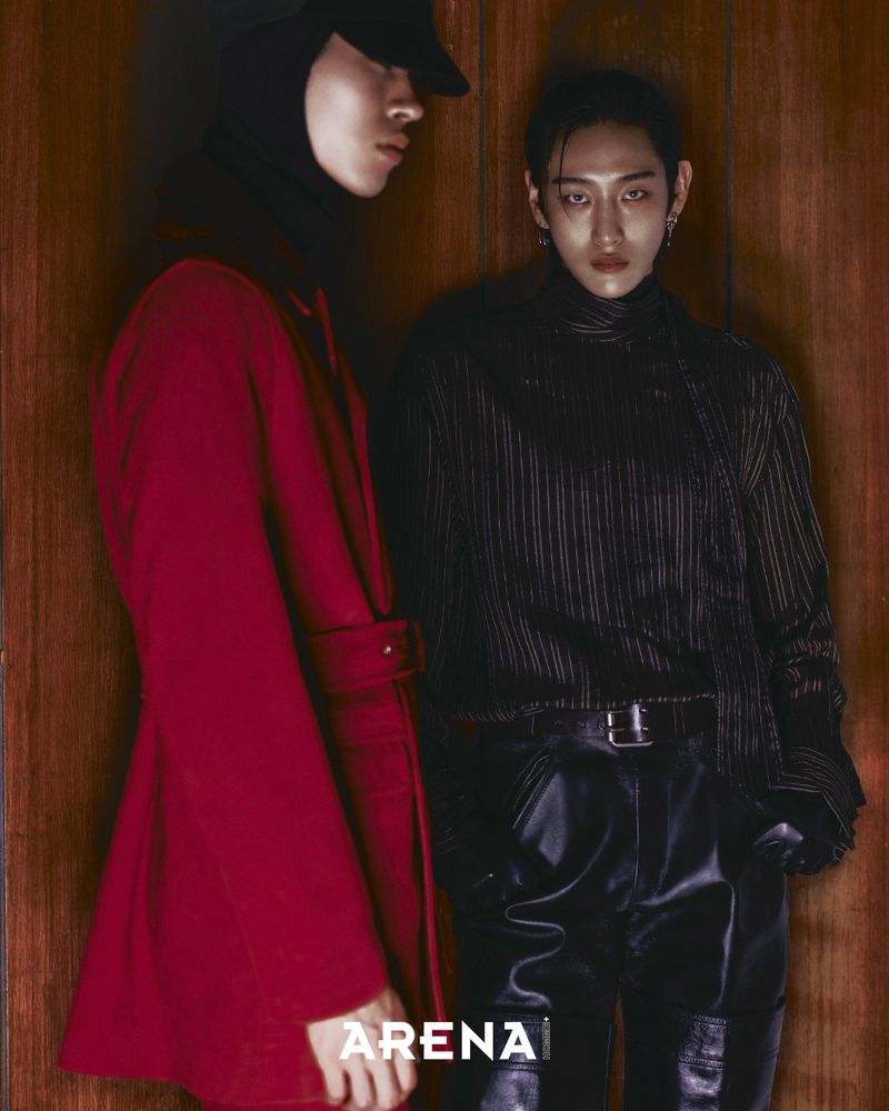 Two androgynous models pose indoors against a wood-panelled wall. One model wears a red coat, and the other is in a black, striped turtleneck.