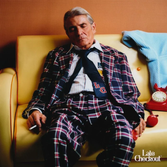 Elderly man in a plaid suit and tie sleeps on a yellow sofa with a cigarette in his hand next to a red rotary phone.