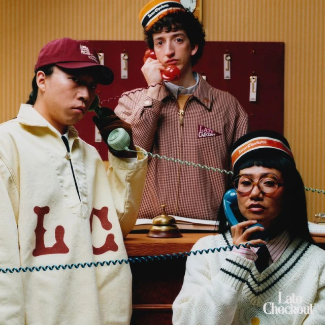 Three people wearing themed uniforms answer vintage telephones in front of a hotel switchboard in a brightly lit indoor scene.