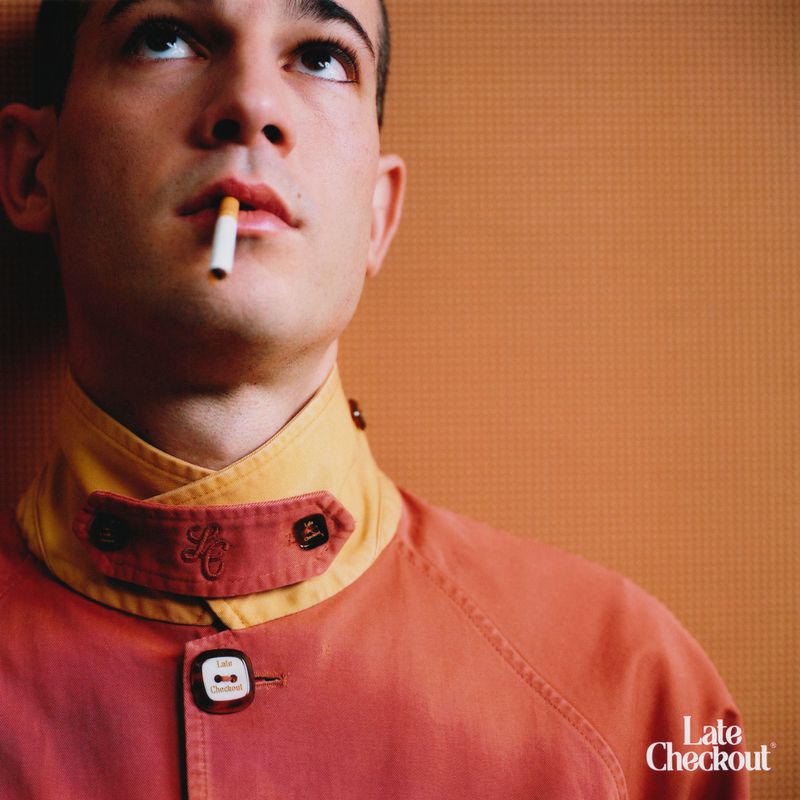 A fashionable young adult stares upward with a cigarette in his mouth, wearing a rust-colored jacket against a peach background.