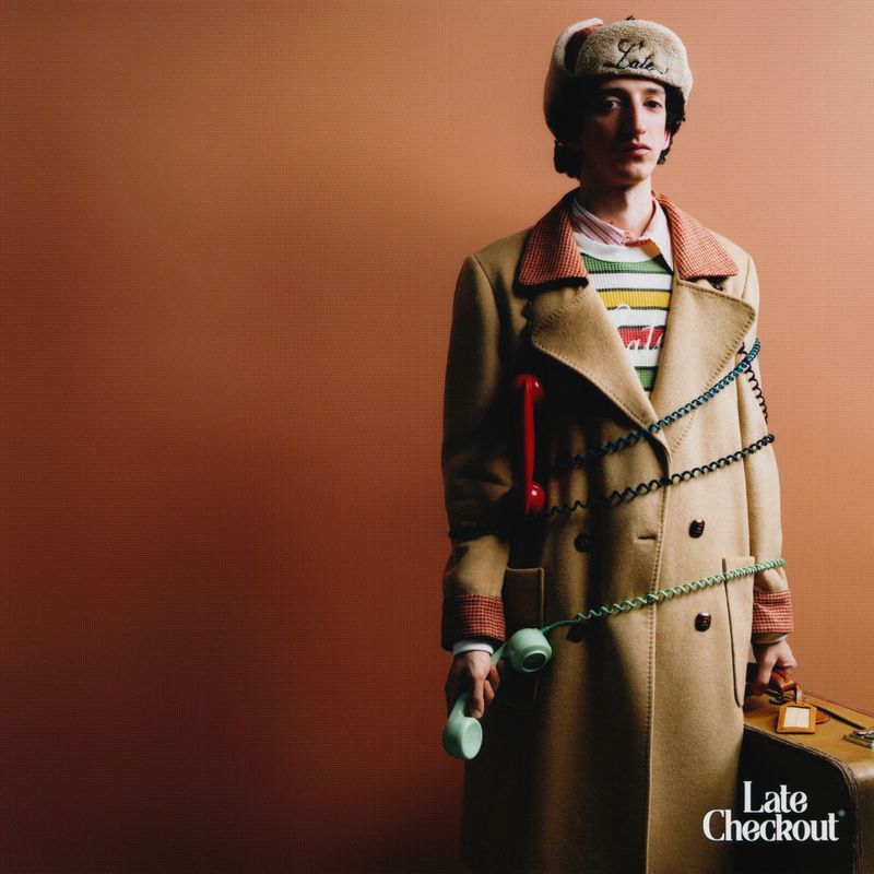 Man in a tan coat and fur hat poses with telephones and a suitcase against a neutral backdrop in a fashion-style portrait.