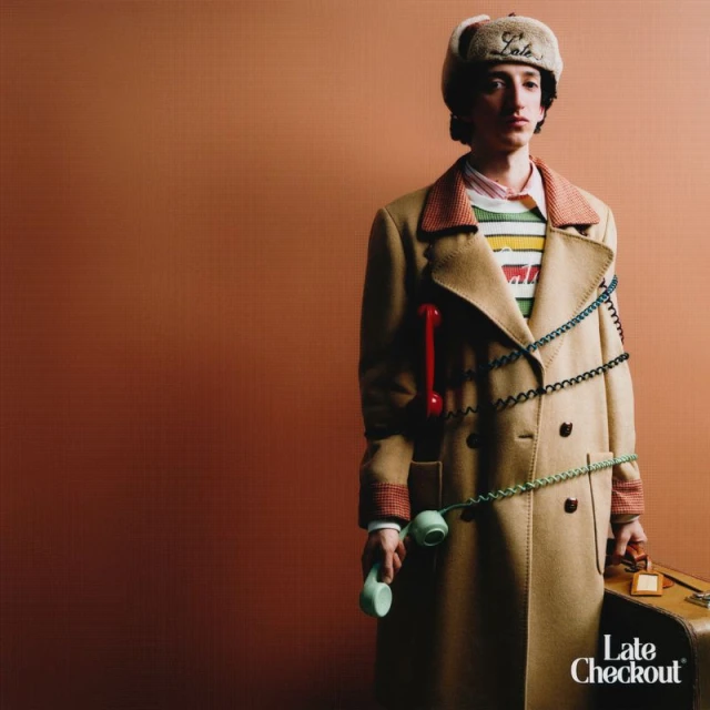 Man in a tan coat and fur hat poses with telephones and a suitcase against a neutral backdrop in a fashion-style portrait.