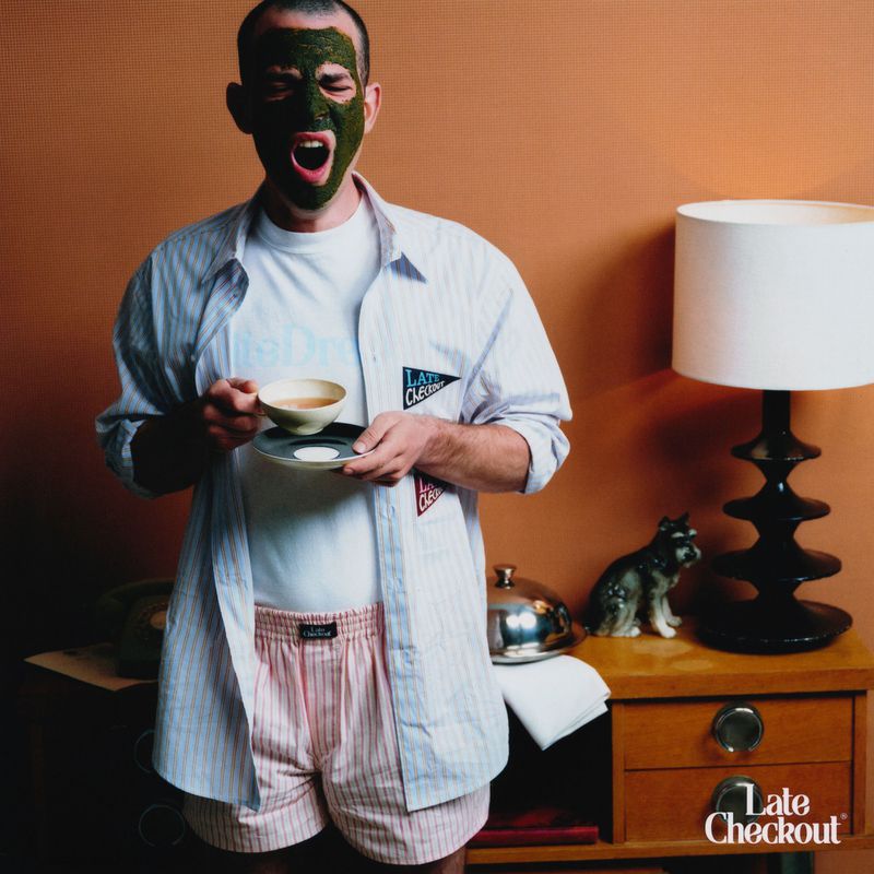 Man with a green face mask, wearing an open striped shirt over a t-shirt and striped boxers, yawning, while holding a bowl and a plate indoors.