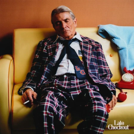 An older man in a navy plaid suit and tie sleeps on a yellow sofa holding a cigarette beside a vintage red rotary telephone.
