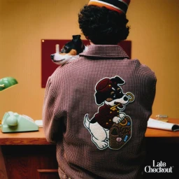 Rear view of a person in a houndstooth jacket with an embroidered dog patch, holding a small dog on their shoulder at a wooden desk.