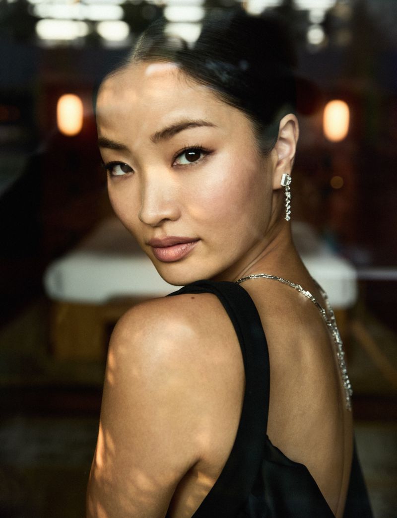 Portrait of a young Asian woman with black hair in formal wear, gazing over her shoulder in a dimly lit indoor setting.