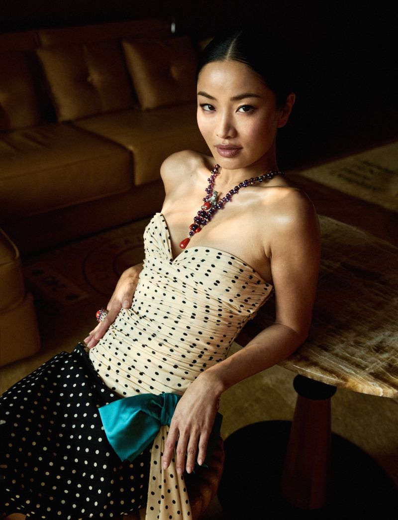 Elegant Asian woman in a polka dot dress and statement necklace leans on a table, with soft lighting and a sofa in the background.