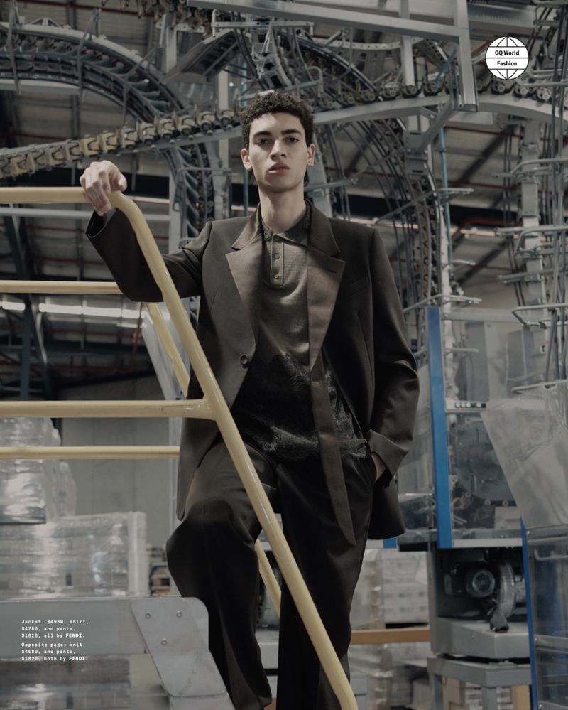 An adult man stands on metal steps in a factory setting, wearing a dark suit with hand in pocket, posing for a fashion editorial.