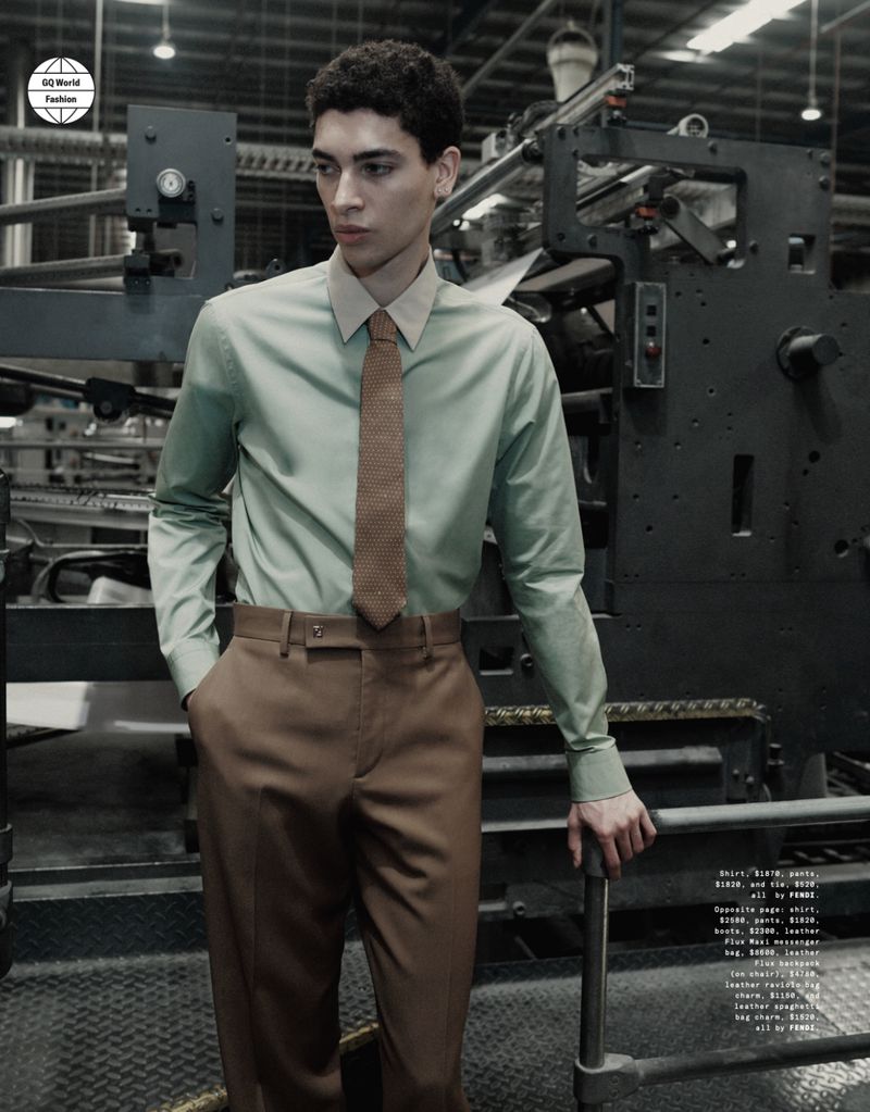 Fashion shoot of a young man in a light green shirt and brown trousers, posing in an industrial setting with machinery in the background.