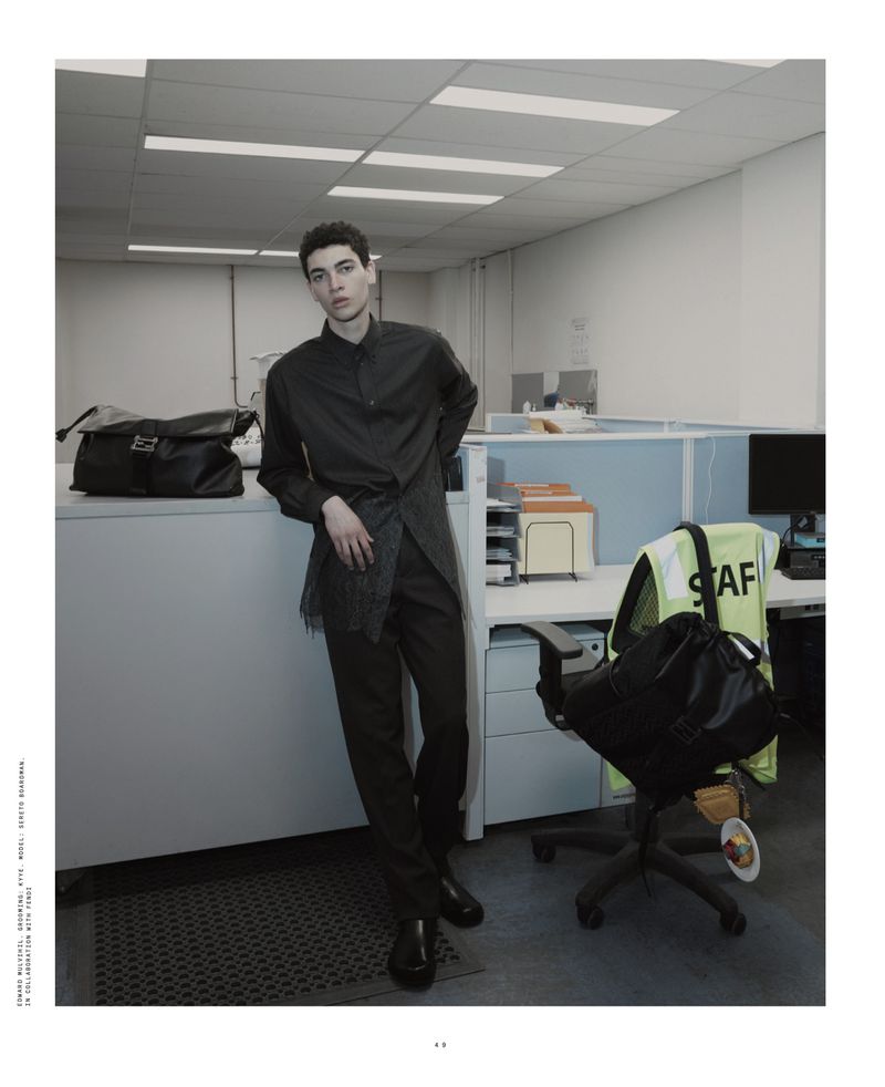 Male fashion model poses in gray button-down shirt and pants near office cubicles, complemented by a large messenger bag and desk chair.