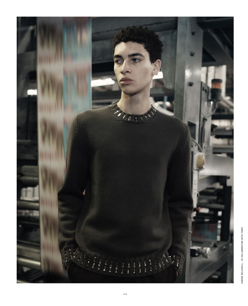An adult male poses in a gray sweater with metallic detailing, with a blurred industrial printing press in the background.