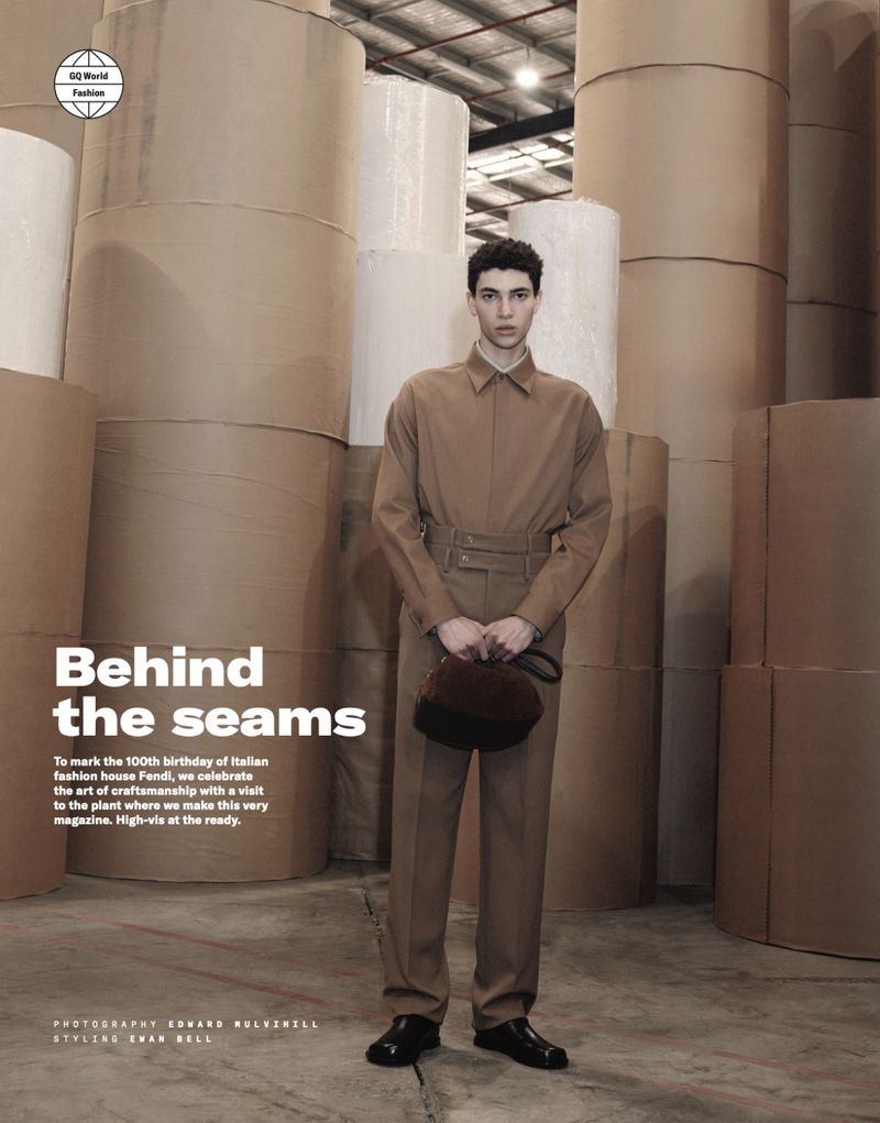 Fashion editorial shot of a young adult male model in tan clothing holding a brown bag, posing in front of rolls of paper.