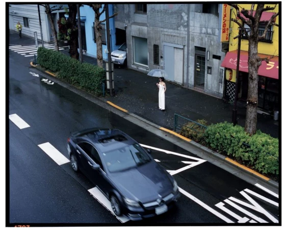 City street scene featuring a woman in a white dress holding an umbrella, with a black car and buildings under overcast daylight.