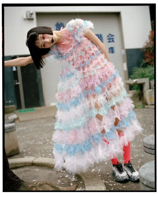 Young woman in a tiered frilly dress stands in front of a building, wearing bright red tights and chunky sneakers in an outdoor setting.