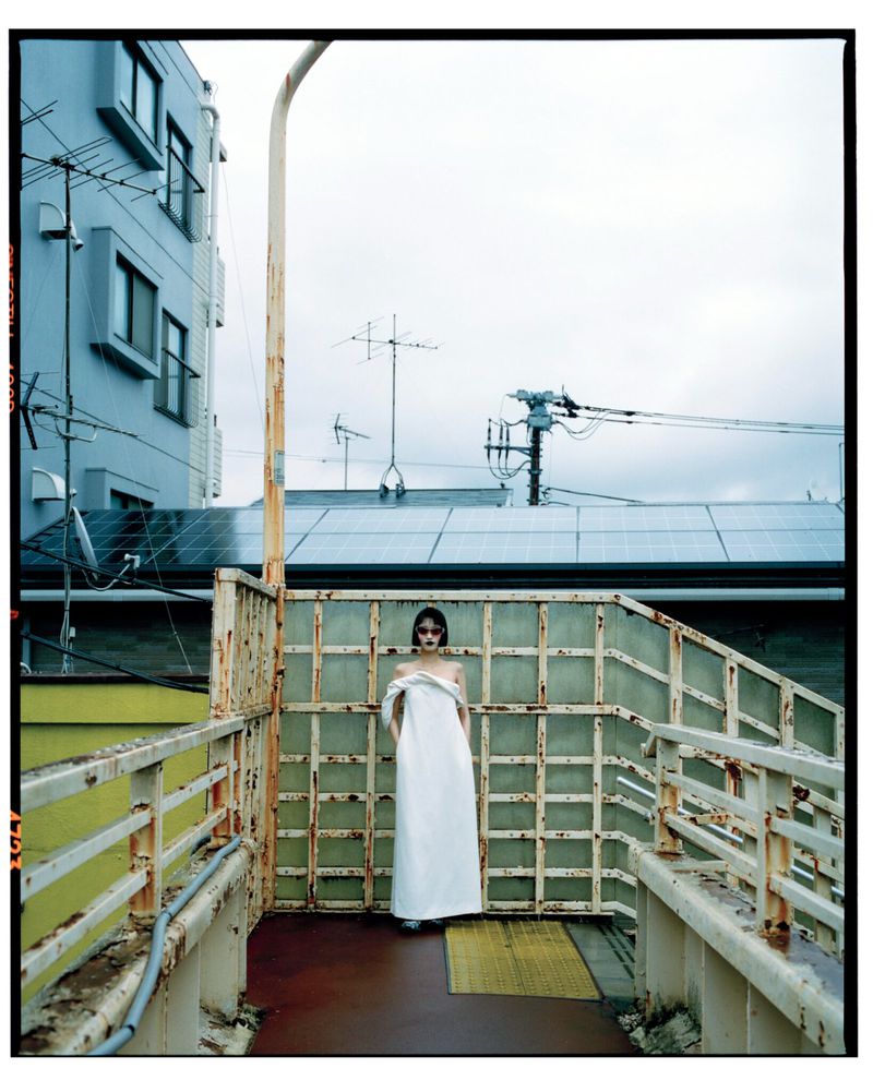 A woman in a long white dress stands on a pedestrian bridge in front of a building, overcast day, fashion editorial photography.