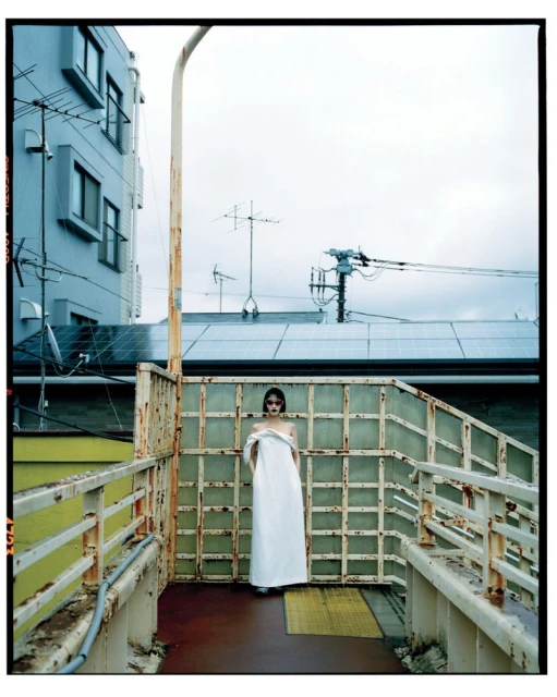 A woman in a long white dress stands on a pedestrian bridge in front of a building, overcast day, fashion editorial photography.