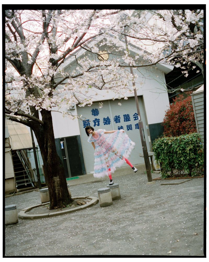 Woman in a colourful dress strikes a pose on concrete blocks beneath a blooming cherry tree with a building in the background.