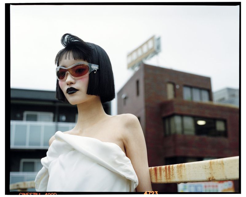 Young woman with bob haircut and sunglasses wearing draped white cloth, posing outdoors with buildings visible in the background.