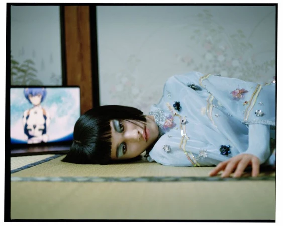 Asian woman poses on a tatami mat floor in a blue beaded dress, with an anime show on a laptop in the background.
