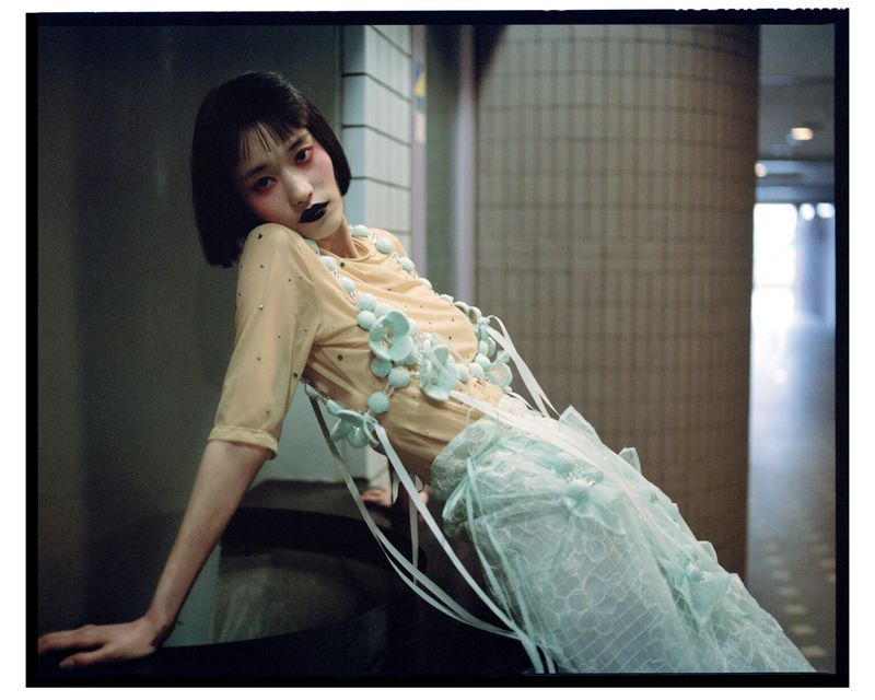 Asian woman with short black hair poses indoors. She wears a peach top, a turquoise skirt, and matching flower accessories.