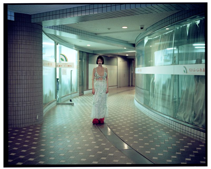 Fashionable young woman in a long white dress and red shoes stands in an empty, tiled corridor with curved glass walls and overhead lighting.